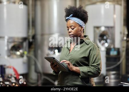 Medium shot of concentrated female African American process technician wearing denim overall taking notes on tablet computer while inspecting tanks wi Stock Photo
