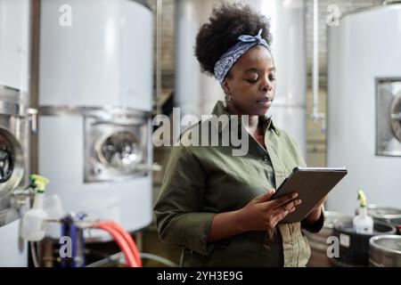 Medium shot of concentrated female African American supervisor using tablet computer inspecting production process in workshop of cider or beer factor Stock Photo