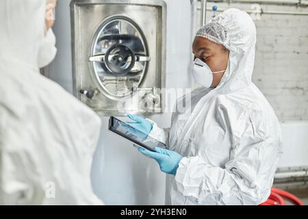 Side view of professional female African American supervisor in mask and hazmat suit using tablet computer while discussing with colleague production Stock Photo