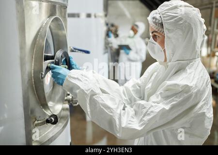 Side view of female process technician wearing hazmat suit and protective gloves turning valve on tank filled with liquid, while inspecting cider ferm Stock Photo