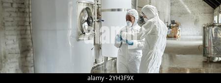 Header image of two professional female supervisors in masks and coveralls using tablet computer discussing production process while inspecting tanks Stock Photo