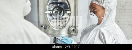 Header image of professional female African American supervisor in mask and hazmat suit using tablet computer while discussing with colleague producti Stock Photo
