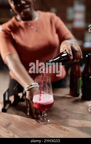 Vertical shot of female African American sommelier pouring craft cider into glass during product sampling in tasting room as part of factory, copy spa Stock Photo