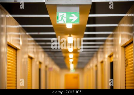 Storage facility hallway with yellow doors, bright lighting, and an overhead green emergency exit sign. Stock Photo