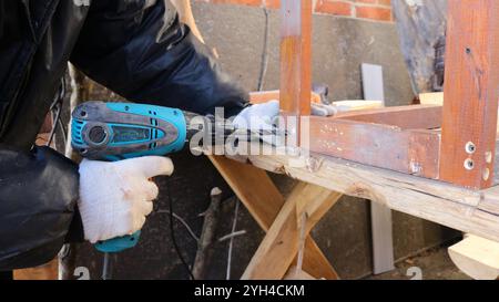 drilling holes in wooden structure using blue drill, carpenter in gloves with hand tool making holes for fasteners in wooden frame Stock Photo