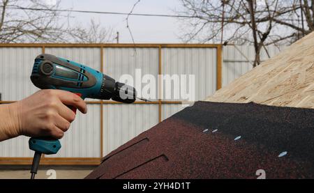 a hand with a screwdriver and a screw at the end is directed towards the roof of a low outbuilding with soft tiles to secure the material Stock Photo