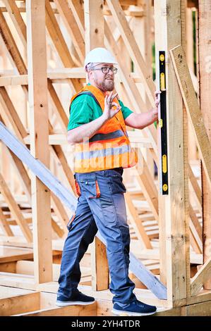Carpenter constructing wooden frame house. Man inspects walls for levelness using spirit level, showing gesture OK sign, wearing protective overalls, helmet and orange vest. Stock Photo