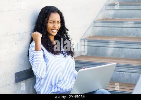 An Excited Young Woman is Joyfully Celebrating Her Success While Using a Laptop on Outdoor Steps Stock Photo