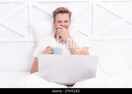 Weekend workaholic. Morning coffee online. Freelancer in bed. Millennial man work on laptop at home. Man lying in bed and surfing internet on laptop. Stock Photo