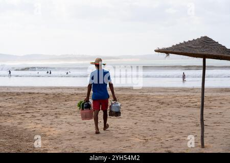 Back view a man carrying a hot teapot and busket with fresh mint walking on the beach selling mint tea. Stock Photo