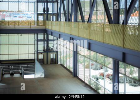 Modern interior with industrial design elements, featuring a glass elevator shaft, yellow metal railing, and large windows overlooking an urban landsc Stock Photo