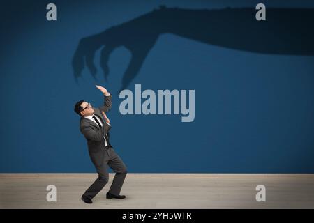 Businessman scared by monstrous hand reaching for him. Creepy shadow on blue wall Stock Photo