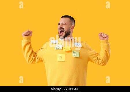 Happy young man and sticky notes with text DAY OFF on his sweatshirt against yellow background Stock Photo