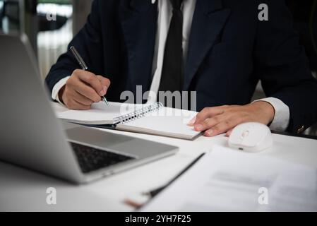 Businessman in a suit writing notes in a notebook with a laptop on a desk in an office setting, focused on work and productivity. Stock Photo