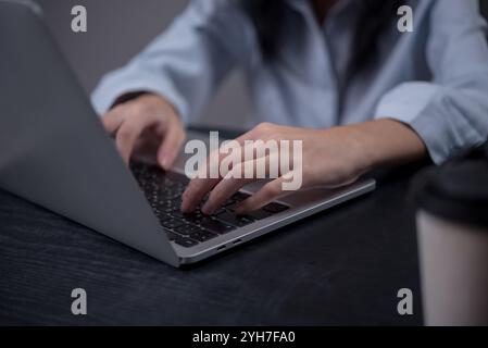 Close-up image of a person's hands typing on a laptop keyboard while sitting at a dark wooden desk with a coffee cup nearby Stock Photo
