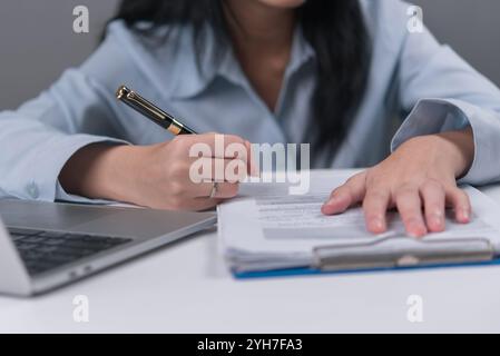 Close-up shot of a woman writing on paperwork using a pen with a laptop next to her on a desk, focusing on the hand and pen Stock Photo