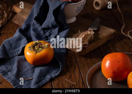 Vibrant persimmons on navy cloth contrast with worn wooden table and vintage kitchenware. Soft lighting emphasizes textures, creating a cozy autumnal Stock Photo