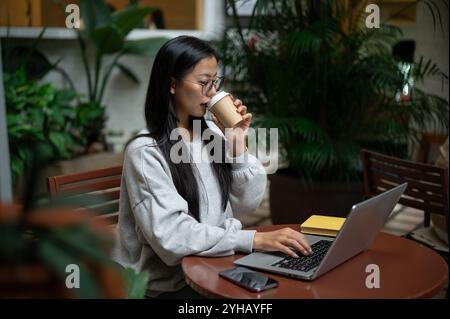 A beautiful, focused Asian woman in a cozy sweater and eyeglasses works remotely from a coffee shop, sipping coffee while working on her laptop at an Stock Photo
