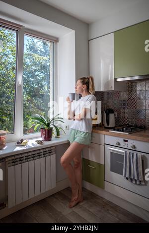 Young relaxed woman enjoying morning coffee or tea standing in kitchen by window with closed eyes. Stock Photo