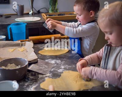 Two kids in aprons make cookies with metal cutters in messy kitchen, focused on learning to make cookies with happy faces and flour-covered hands as Stock Photo