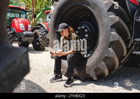 Female farm worker sitting on tractor wheel, using digital tablet at agricultural equipment dealership. Stock Photo
