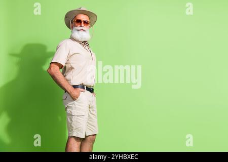 Elderly man in safari-style outfit posing confidently against a vibrant green background, embodying adventurous spirit and wanderlust. Stock Photo