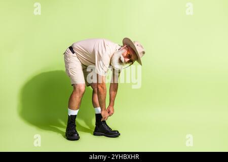 Elderly man in safari outfit preparing for an adventurous journey, tying hiking boots, set against a vibrant green backdrop Stock Photo