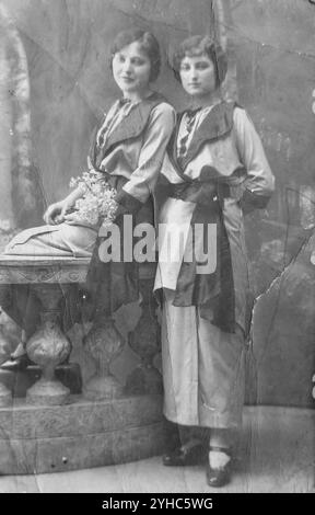 Novi Sad, Yugoslavia - 1920s: Portrait of two cute stylish young women, one sitting on a beautiful marble fence, other one standing next to her, with wave hairstyle, wearing long bright dresses with wide black collars and waist sash and wrist corsage Stock Photo