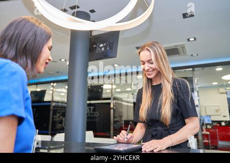 One woman talks to another at a counter, filling in on the tablet. The woman on the left is wearing a blue shirt Stock Photo
