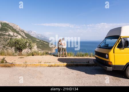 Couple enjoying the view of Croatian mountainous coast standing by their retro yellow camper van parked on a cliff, Croatia Stock Photo