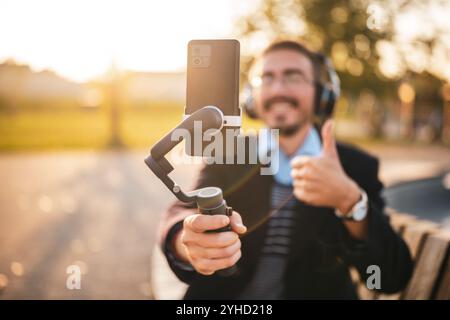 Businessman vlogger with headphones using mobile phone with gimbal.He is showing thumb up while recording vlog outdoor. Focus on the phone and gimbal. Stock Photo
