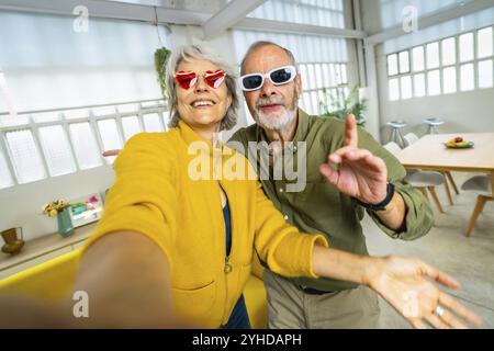 Senior woman with heart shape sunglasses taking selfie with husband while having fun at home Stock Photo