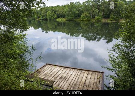 A tranquil lake with a wooden jetty surrounded by a dense forest that is reflected in the water Stock Photo