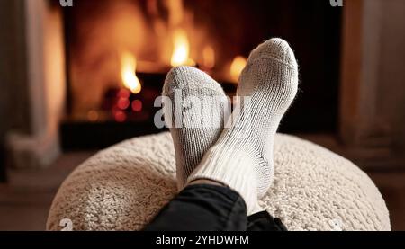 Cozy Adult feet in front of fire in light beige oatmeal color warm wool knitted socks, resting on a neutral white boucle fabric footstool in lounge li Stock Photo