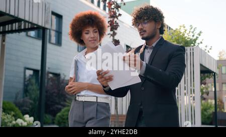 Indian Arabian male man businessman holding papers documents corporate discuss result plans with African American woman colleague businesswoman Stock Photo