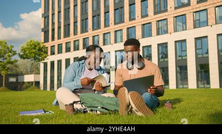 Two friends African American ethnic students sitting on green grass meadow lawn in park outdoors woman man girl guy doing homework university prepare Stock Photo