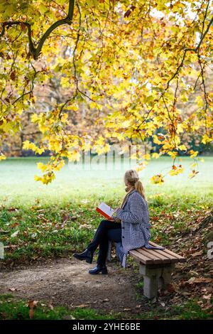 Woman in houndstooth coat sitting on bench and reading book in autumn park. Relaxation outdoors Stock Photo
