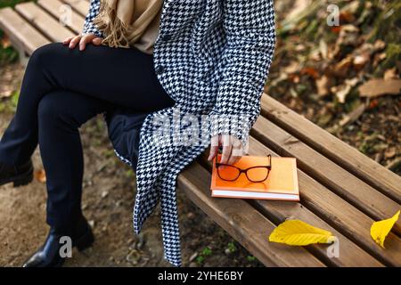 Reading book in autumn park. Woman in houndstooth coat sitting on bench and holding eyeglasses Stock Photo