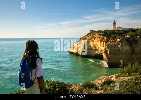 A young girl views the Alfanzina Lighthouse from a hiking trail on a sunny day, framed by Algarve’s rugged cliffs and turquoise ocean Stock Photo