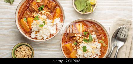 Two bowls of delicious massaman curry with beef, potatoes, rice, and peanuts, garnished with parsley on white wooden background, Thai cuisine Stock Photo