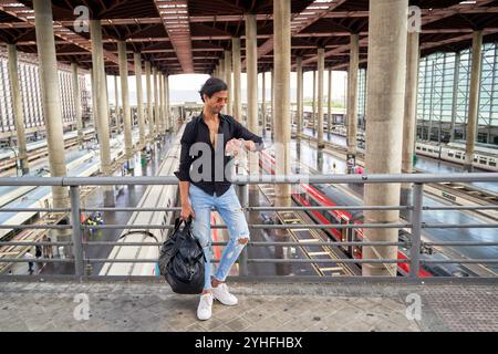 Indian man in black shirt and ripped jeans checking his watch on a train platform bridge, holding a black bag. Stock Photo
