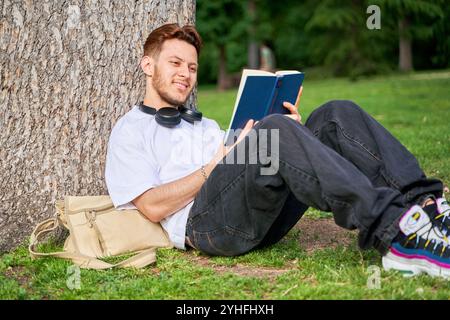 A man is sitting on the grass reading a book. He is wearing headphones and has a backpack next to him. The scene is peaceful and relaxing, with the ma Stock Photo