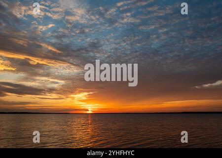 Fiery orange and yellow clouds filling the sky over the calm water of a lake as the sun sets on the distant horizon. Stock Photo
