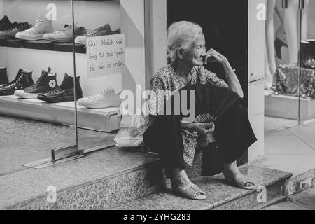 An elderly woman, seated on the steps of a store with a fan in hand, takes a moment of rest while the hustle of the street continues around her. Stock Photo