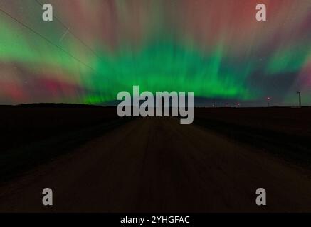 The aurora borealis glows above a quiet country road, with a lone tree standing by the roadside, windmills in the distance, and a satellite crossing t Stock Photo