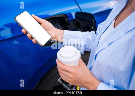 Woman charges their electric car, holding a coffee cup and checking their smartphone nearby Stock Photo