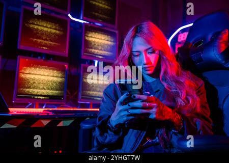 Asian woman in front of a computer monitor display using mobile phone, neon light, Hacker wear black Stock Photo