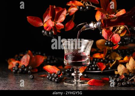 Black chokeberry drink is poured from a bottle into a glass. Sweet drink and fresh berries on a black stone table. Stock Photo