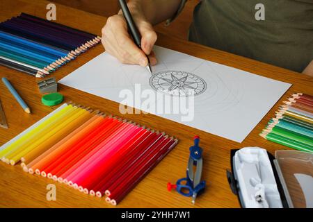 Hand of a Caucasian woman drawing a mandala pattern on a white paper lying on a wooden table, colorful pencils and other drawing utensils nearby Stock Photo