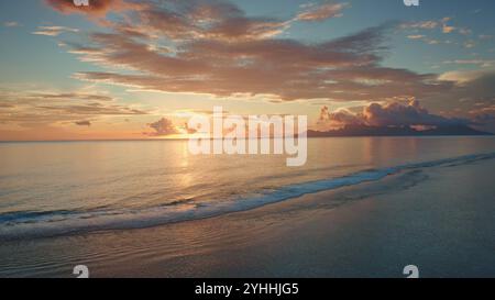 Golden sunset illuminate tranquil ocean, Moorea island in background, tropical paradise scene in French Polynesia. Colorful orange cloudy sky reflect in sea waves. Exotic summer travel. Aerial flight Stock Photo